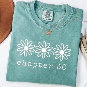 May include: A sage green short-sleeve t-shirt featuring a white graphic of three daisy flowers arranged in a horizontal row above the text "chapter 50" in a white serif font. The shirt has a crew neckline and a Comfort Colors brand label.