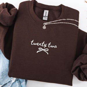 May include: A dark brown sweatshirt with the text "twenty two" in white script and a bow graphic. A silver necklace with a circular pendant is draped across the sweatshirt. The sweatshirt is folded, with a portion of blue jeans visible.