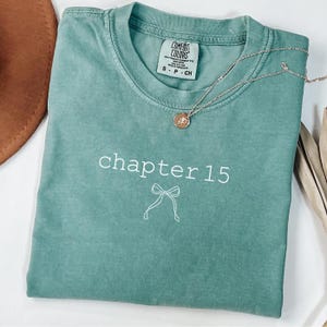 May include: Seafoam green Comfort Colors t-shirt with the text "chapter 15" and a bow graphic. A silver necklace with a pendant rests on the shirt. The shirt is folded and has a tag that reads "S - P - CH".