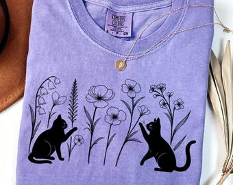 May include: A lavender short-sleeve t-shirt featuring a black line art graphic of two cats sitting among various wildflowers. The shirt has a crew neckline and a Comfort Colors brand label printed on the inside of the collar area.