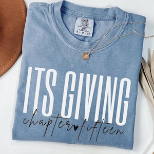 May include: A blue Comfort Colors t-shirt with the text "ITS GIVING chapter fifteen" in white and black script. A gold necklace with a pendant is draped across the shirt. The shirt is folded and has a small tag.