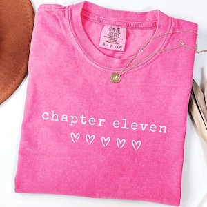 May include: A bright pink t-shirt with the words "chapter eleven" and five heart symbols printed in white. A gold-colored necklace with a circular pendant rests on the shirt. The shirt has a Comfort Colors tag.