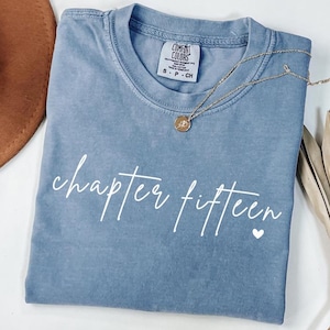 May include: A blue Comfort Colors t-shirt with the words "chapter fifteen" in white script and a small white heart. A gold necklace with a pendant rests on the shirt. The shirt has a small tag that reads "Comfort Colors".