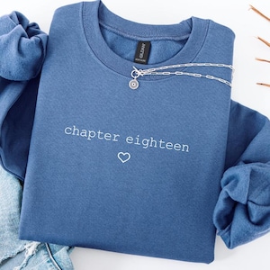 May include: A blue sweatshirt with the text "chapter eighteen" and a heart symbol. A silver necklace with a sunburst pendant is draped across the sweatshirt. The sweatshirt is folded with a denim garment visible.