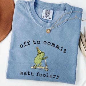 May include: A light blue t-shirt featuring a graphic of a green frog wearing a party hat and riding a scooter. The text "off to commit math foolery" is printed in black letters above and below the frog. A gold-toned necklace rests on the collar.