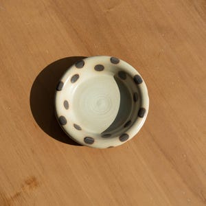 May include: A small, off-white ceramic bowl with dark brown polka dots around the rim. The interior of the bowl has a light green hue and a spiral pattern. The bowl is on a wooden surface.
