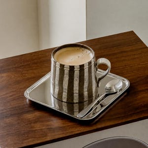 May include: A ceramic mug with a dark brown and white vertical striped pattern filled with a light brown coffee beverage. The mug sits on a square, polished silver-toned metal tray alongside a matching silver-toned metal spoon.