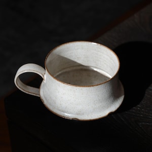 May include: A speckled, off-white ceramic mug with a rounded base and a curved handle. The rim and base edge have a brown accent. The mug sits on a dark wooden surface, with shadows adding depth.