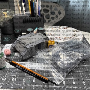 May include: A gray plastic miniature tank model sits on a cutting mat alongside a clear plastic bag containing additional black plastic parts, a paint palette with dried paint, a small yellow paint bottle, and a black brush with an orange handle.