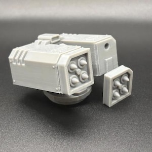 Puede incluir: Modelo gris claro impreso en 3D de un sistema de armas de múltiples cañones. El modelo presenta múltiples componentes rectangulares con proyectiles cónicos que sobresalen. El diseño sugiere un tema futurista o de ciencia ficción.