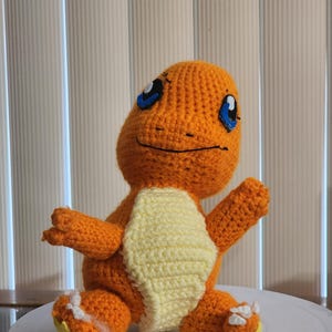 May include: A handmade orange crochet Charmander plush toy with blue eyes and a cream-colored belly. The plush is sitting with its arms and legs outstretched. The toy is a popular character from the Pokemon franchise.