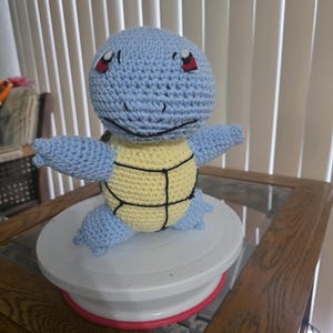 May include: A handmade crochet Squirtle plush toy. The toy features a light blue head, arms, and legs, with a yellow shell. The eyes are red and the mouth is black. The plush is sitting on a white cake stand.