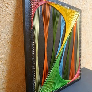 Könnte beinhalten: Ein quadratisches String-Art-Stück mit einem schwarzen Holzrahmen. Das Kunstwerk zeigt ein farbenfrohes Design aus orangefarbenen, gelben, grünen, blauen und rosa Fäden, die über Nägel gespannt sind und ein geometrisches Muster bilden. Das Stück wird vor einem neutralen Hintergrund präsentiert.
