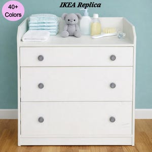 May include: A white three-drawer changing table with gray knobs. The top surface holds baby supplies, including diapers, a plush bear, and bottles. The text "IKEA Replica" is at the top. A pink circle with the text "40+ Colors" is in the upper left corner.