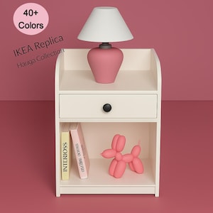 Puede incluir: Una mesita de noche blanca con un cajón y un estante inferior. Una lámpara rosa con una pantalla blanca está encima. Un perro globo rosa y dos libros están en el estante. El texto "40+ Colors" y "IKEA Replica Häuga Collection" es visible.