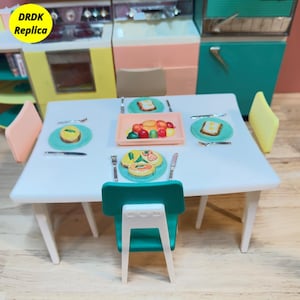 Puede incluir: Una mesa de comedor blanca en miniatura para cuatro personas con platos turquesa, una bandeja de fruta y sillas turquesa y melocotón. La mesa está puesta con comida de juguete, cuchillos y tenedores. El texto "DRDK Replica" está en la esquina superior izquierda.