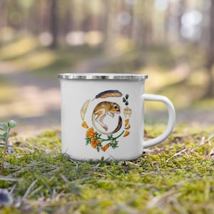 May include: White enamel mug with a silver rim, featuring a watercolour illustration of a small, brown and tan animal surrounded by feathers, flowers, and other natural elements. The mug has a white handle and is set against a blurred outdoor background.