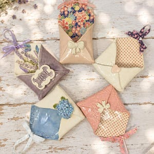 May include: Five ceramic envelope-shaped ornaments with hanging ribbons. One is purple with lavender, one is peach with floral patterns, one is cream with polka dots, one is blue with blue flowers, and one is pink with embossed patterns and polka dots.