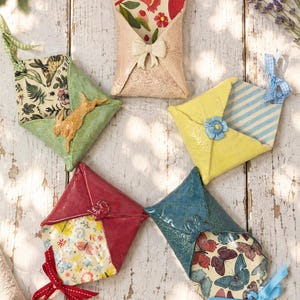 May include: Five ceramic envelope-shaped ornaments with hanging ribbons. Each features a unique glazed exterior, including floral, butterfly, and rabbit patterns, with decorative accents like bows and flowers attached to the front of each piece.