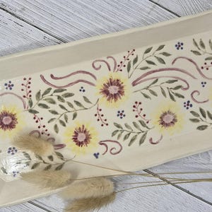 May include: Cream-coloured rectangular ceramic serving platter. It features yellow sunflowers, green leaves, and burgundy accents. The edges have a scalloped design. Measurements are in centimetres.