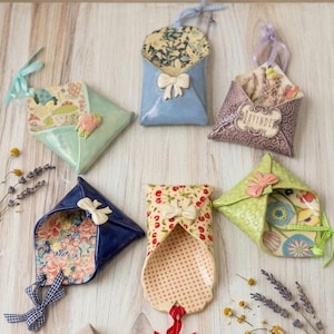 May include: Six ceramic envelope-shaped wall pockets in various colors and patterns, including floral, polka dot, and solid glazes. Each piece features a small sculpted bow and a ribbon loop for hanging, with one labeled Lavender.