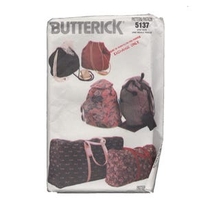 May include: A Butterick 5137 sewing pattern envelope featuring illustrations of various bags, including drawstring backpacks and duffel bags in black, red, and floral patterns. The white paper envelope displays the brand name and pattern number.
