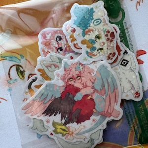 May include: A collection of colorful, die-cut stickers featuring whimsical, fantasy-themed illustrations. The stickers showcase various creatures with wings, floral accents, and a soft, pastel color palette. The stickers are packaged in a clear plastic sleeve.