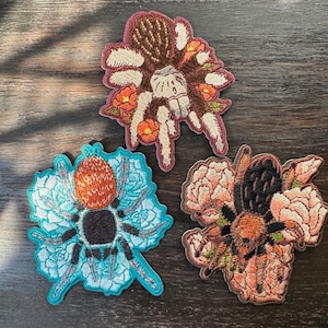 May include: Three embroidered iron-on patches featuring a spider with floral accents. The patches are in shades of brown, blue, and pink.