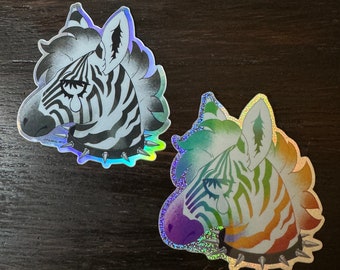 May include: Two iridescent holographic stickers of zebra heads with a punk rock aesthetic. The zebras have black and white stripes and are wearing studded collars. The stickers are on a brown wooden surface.