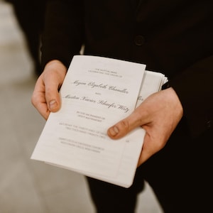 May include: A person holding a stack of white wedding programs. The programs have black text with the names "Megan Elizabeth Chandler" and "Martin Xavier Schaefer Thirty" visible. The person is wearing a black suit.