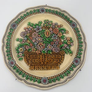 May include: A round ceramic decorative plate with a scalloped edge featuring a raised relief design of a brown woven basket filled with a colorful assortment of flowers in shades of pink, yellow, blue, and green, surrounded by a floral border.