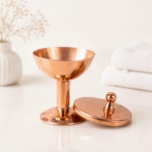 May include: A polished copper lidded container with a wide, shallow bowl, a cylindrical stem, and a circular base. The matching circular lid features a small, rounded knob handle. The metal surface has a warm, reflective, metallic finish.