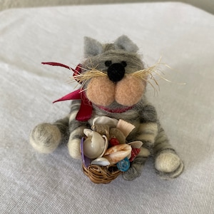 May include: A gray and white striped cat with a red ribbon around its neck holds a basket full of seashells. The cat has a black nose and brown eyes.