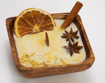May include: A square wooden bowl candle with a creamy wax base. The candle is decorated with a dried orange slice, a cinnamon stick, star anise, and dried flower petals. The candle is a warm, inviting home decor item.