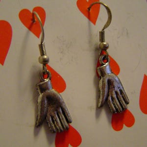May include: A pair of silver-toned hand-shaped earrings hanging in front of a playing card. The card is the six of hearts, with red heart symbols. The background includes a white surface with red lipstick kiss marks.