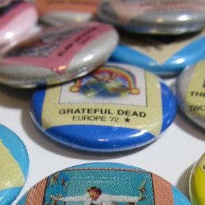 May include: A collection of vintage music-themed pinback buttons. The buttons feature album art and text from artists like Elton John, Eagles, and Grateful Dead. The buttons are various colors, including blue, yellow, and pink.