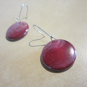 May include: A pair of drop earrings featuring circular, deep red, glossy pendants with a subtle textured finish. Each pendant is attached to a thin, silver-toned metal hook wire, allowing the round pieces to hang freely from the ear wire attachment.