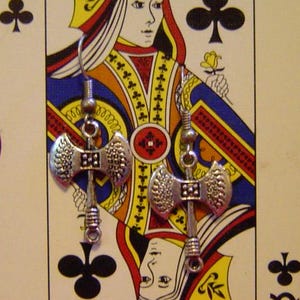 May include: Silver-colored axe-shaped earrings hanging in front of a Queen of Clubs playing card. The card is on a red surface, with a white object decorated with red and pink lip prints in the background.