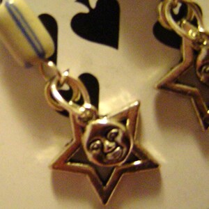 May include: Gold-toned earrings featuring star-shaped charms with a smiling face design. Each earring has a blue and yellow striped bead. The earrings are displayed on a playing card with black spade symbols.