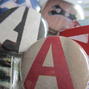 May include: A collection of colorful button pins with various designs. One pin features a large red "A" on a tan background. Other pins display abstract patterns and graphic elements in red, black, white, and blue. The pins are round and have a glossy finish.