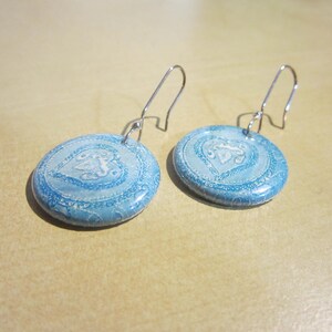 May include: A pair of round, light blue dangle earrings featuring a white heart-shaped pattern with intricate lace-like detailing. The earrings are suspended from silver-toned metal ear wires, showcasing a smooth, glossy finish on the circular discs.