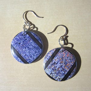 May include: A pair of round dangle earrings. Each earring features a blue and white patterned design with black diagonal stripes. The earrings have silver-colored hooks.