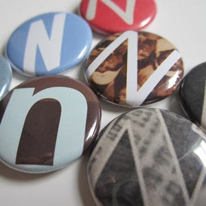 May include: A collection of round buttons with the letter "N" or "n" in various colors and patterns. The buttons feature a white letter on backgrounds of blue, red, brown, and patterned designs. The buttons are glossy.