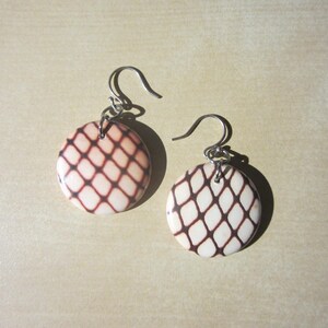 May include: A pair of round earrings with a cream-colored base and a brown net-like pattern. The earrings have silver-colored hooks. The earrings are displayed on a light beige surface.