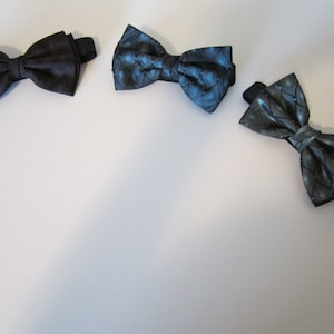 May include: Three bow ties in shades of black and blue are displayed against a white background. The bow ties have different patterns, including a solid black, a blue with a subtle pattern, and a gray with a diamond pattern. Each bow tie has a black adjustable strap.