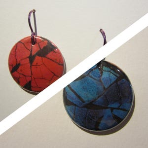 May include: A pair of round earrings with a cracked glass design. One earring is red with black cracks, the other is blue with black cracks. Both have purple ear wires.
