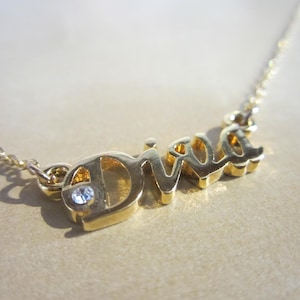 May include: A gold-colored necklace with the word "Diva" spelled out in a cursive font. The letter "D" has a small, round, clear gemstone. The necklace chain is delicate and gold-colored.