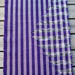 May include: A purple and white striped rug with a woven design in the upper right corner. The rug has a fringe border.