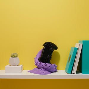 May include: A black video game controller rests on a purple octopus-shaped stand. The stand is on a white shelf with a small succulent in a geometric white pot and a stack of books. The background is yellow.