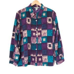 May include: A long-sleeved button-down shirt with a geometric pattern in teal, purple, and beige squares. The shirt is hanging on a wooden hanger against a white background. The collar is pointed and the buttons are white.